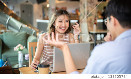 Asian woman smiling as a man talking while sitting with a laptop at wooden table in cafe or canteen. 135305784