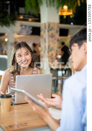 Asian woman looking at a man holding tablet talking as sits with laptop at table in cafe or canteen 135305788