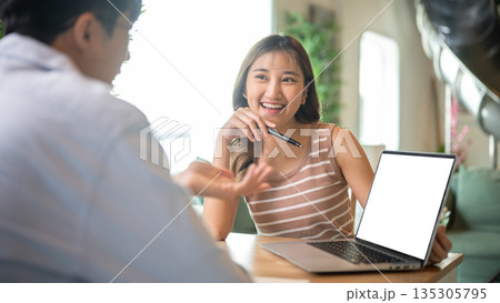 Asian woman holding pen talking showing white screen laptop to friend as sitting in cafe or canteen. 135305795