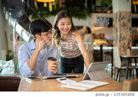 Asian woman standing and bending over wooden table looking at a man's tablet in cafe or canteen. 135305808