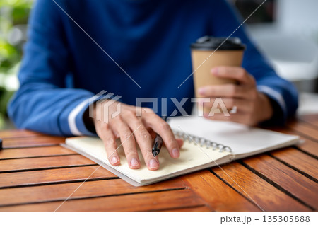 Close up of hand with pen and other holding coffee over notebook at wood table in cafe or canteen. 135305888