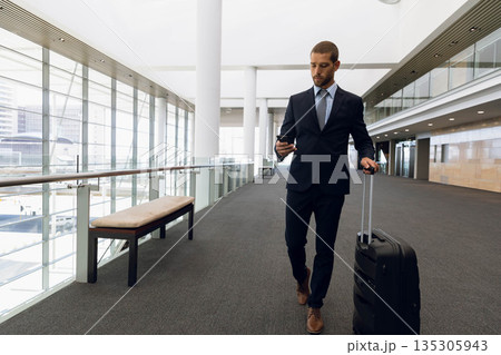 Male business traveler walking at airport pulling suitcase and checking smartphone, copy space 135305943