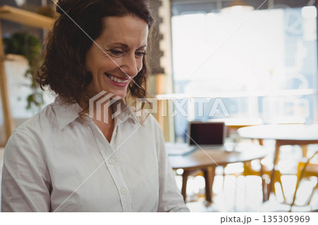 Middle-aged woman leaning over wooden table at co-working cafe, reviewing paperwork beside laptop 135305969