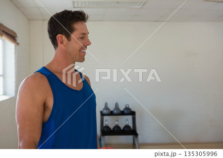 Man standing in gym wearing blue tank top beside kettlebell rack and window blinds, copy space Man standing in gym wearing blue tank top beside kettlebell rack and window blinds, copy space 135305996