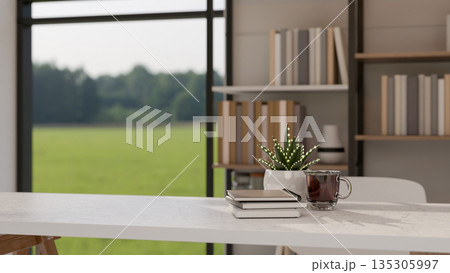 Glass of coffee and book aside pot plant on white table in a bright living room with greenery view. Glass of coffee and book aside pot plant on white table in a bright living room with greenery view. 135305997