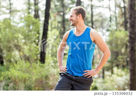 Man in sportswear standing hands on hips, smiling and looking left in forest clearing, copy space 135306032