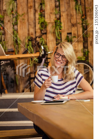 Sitting at communal table holding coffee cup at rustic cafe with pastry and tablet, copy space 135306043