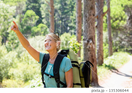 Senior woman pointing and smiling on forest trail wearing backpack with rolled mat and sunglasses 135306044
