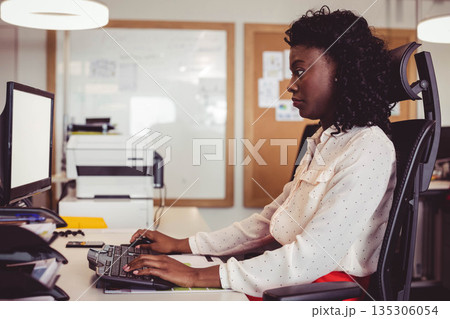 African American woman typing at office desk in office attire with keyboard, monitor, copy space 135306054