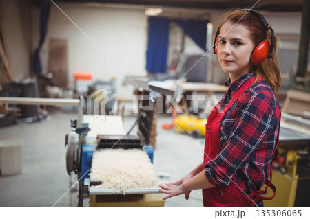 Female carpenter with red apron and earmuffs using planer in workshop making shavings, copy space Female carpenter with red apron and earmuffs using planer in workshop making shavings, copy space 135306065