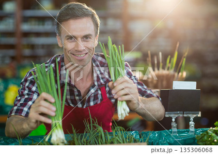 Mature man vendor smiling behind plastic-covered table holding green onions in grocery store Mature man vendor smiling behind plastic-covered table holding green onions in grocery store 135306068