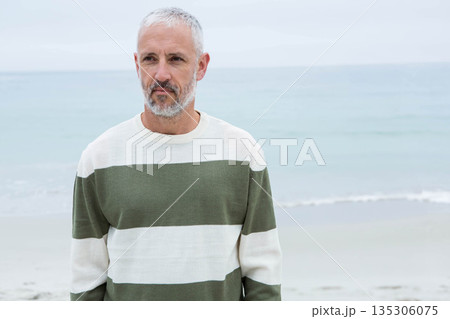 Senior man standing on sandy shoreline at beach wearing olive green striped crewneck sweater 135306075