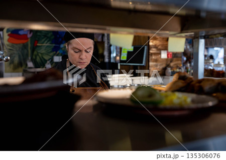 Senior chef wearing jacket arranging plates with poultry garnishes on kitchen pass-through shelf 135306076