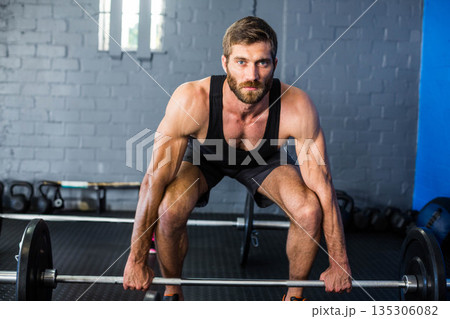 Mid adult male wearing black tank top and shorts lifting loaded barbell at gym with kettlebells 135306082