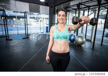 Woman wearing teal sports bra and black leggings lifting dumbbells near exercise balls inside gym 135306088