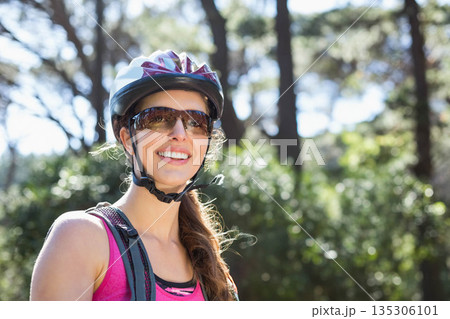 Woman in twenties pedaling on forest trail wearing tank top, helmet, sunglasses and hydration pack Woman in twenties pedaling on forest trail wearing tank top, helmet, sunglasses and hydration pack 135306101
