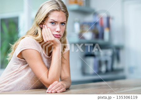Adult woman leaning on wooden countertop and resting cheek on hand near shelving in home kitchen 135306119