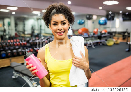 African American woman holding pink water bottle and white towel wearing yellow top at gym 135306125