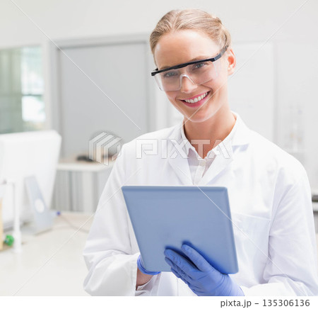 Female lab technician wearing white coat, safety goggles, gloves, holding tablet among lab benches 135306136