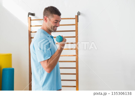 Man lifting hex dumbbell by stall bars with foam rollers in rehab room wearing polo shirt 135306138