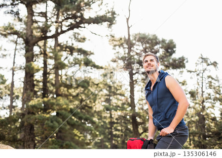 Man kneeling on rocky outcrop in pine forest wearing navy hoodie gripping red backpack, copy space 135306146