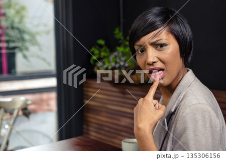Asian woman in gray blazer holding white mug sitting at cafe table touching lip, copy space 135306156