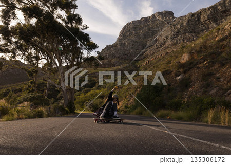 Female skateboarder crouching low on longboard under white helmet gliding down mountain road Female skateboarder crouching low on longboard under white helmet gliding down mountain road 135306172