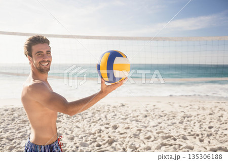 Athletic male standing on sandy beach holding volleyball near net under clear sky, copy space 135306188