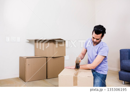 Male kneeling sealing box with packing tape next to blue couch at room corner, copy space Male kneeling sealing box with packing tape next to blue couch at room corner, copy space 135306195