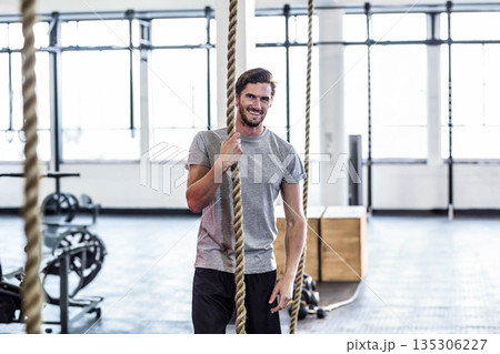 Man grasping thick climbing rope inside gym with weight racks wearing gray athletic tee and shorts 135306227