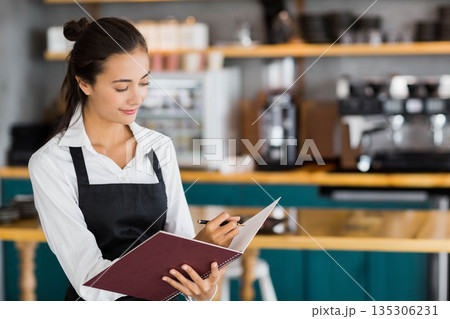 Female barista writing order notebook at cafe counter wearing apron with espresso machine and pen Female barista writing order notebook at cafe counter wearing apron with espresso machine and pen 135306231