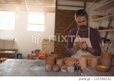 Mid adult male potter wearing apron applying glaze on clay pot at workshop workbench, copy space Mid adult male potter wearing apron applying glaze on clay pot at workshop workbench, copy space 135306232