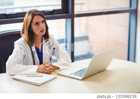 Woman doctor wearing scrubs and stethoscope sitting at desk reviewing notes on laptop and notebook 135306239