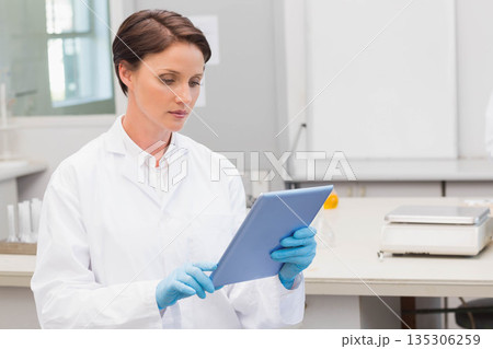 Gloved woman scientist in lab coat reviewing data on tablet at bench with balance, copy space 135306259