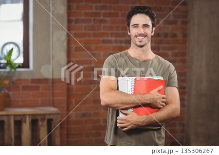 Male student smiling while holding spiral notebooks and red folder in brick-walled room with plant 135306267