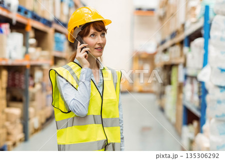 Mid adult woman wearing safety vest and yellow hard hat talking on smartphone inspecting warehouse 135306292