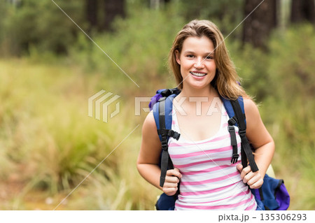 Smiling female hiker holding backpack straps while standing in forest clearing among tall grass 135306293