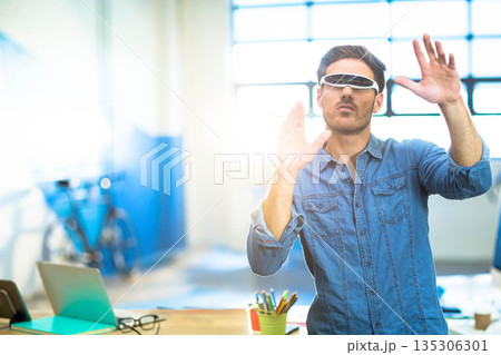Adult man standing behind wooden desk at modern studio wearing white VR glasses and using laptop Adult man standing behind wooden desk at modern studio wearing white VR glasses and using laptop 135306301