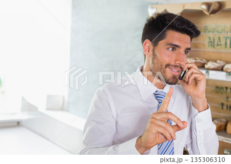 Male bakery manager holding smartphone and gesturing behind counter with bread on shelves in bakery Male bakery manager holding smartphone and gesturing behind counter with bread on shelves in bakery 135306310
