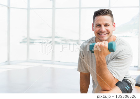 Man kneeling on gym floor wearing gray t-shirt holding teal dumbbell in bright studio, copy space Man kneeling on gym floor wearing gray t-shirt holding teal dumbbell in bright studio, copy space 135306326