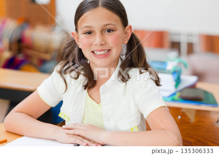 Girl student sitting at wooden desk in classroom smiling with hands folded on notebook and binder 135306333