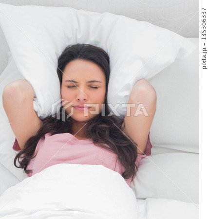 Woman holding pillow against ears while lying under white blanket in bedroom with quilted headboard Woman holding pillow against ears while lying under white blanket in bedroom with quilted headboard 135306337
