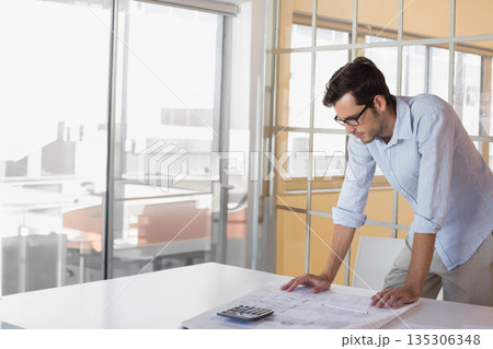 Adult male professional leaning over table studying blueprints and using calculator in meeting room 135306348