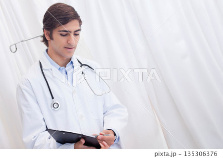 Male doctor in lab coat writing patient notes in clinic with clipboard and stethoscope, copy space 135306376