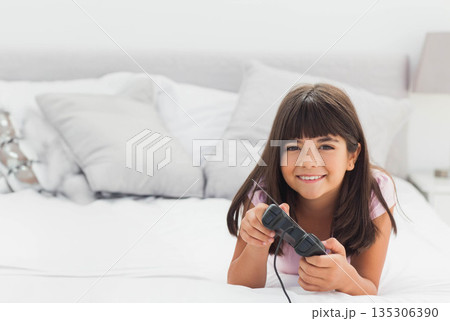 Girl lying on bed holding black wired game controller among gray and white pillows in bedroom Girl lying on bed holding black wired game controller among gray and white pillows in bedroom 135306390