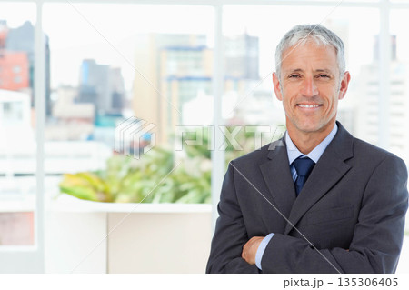 Middle-aged male standing with crossed arms in office by window panes with planter, copy space 135306405