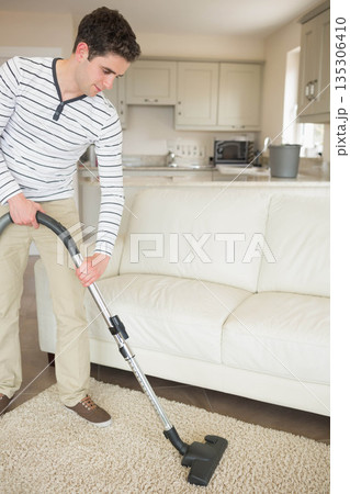 Man vacuuming shag rug near leather sofa and microwave-equipped kitchen with plant, copy space 135306410