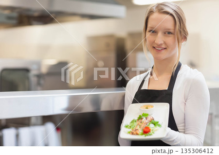 Woman wearing black apron holding salad platter and sauce ramekin inside kitchen, copy space Woman wearing black apron holding salad platter and sauce ramekin inside kitchen, copy space 135306412