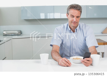 Middle-aged man holding smartphone and eating cereal at kitchen counter near white mugs and toaster 135306415