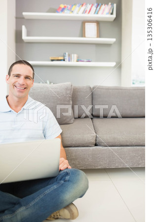 Man in his thirties sitting cross-legged in living room by gray couch holding silver laptop 135306416